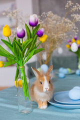 Little red decorative rabbit on a table decorated for Easter with eggs and a bouquet of tulips.