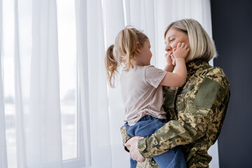 little girl with ponytails touching face of smiling mom in military uniform near window.