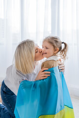 blonde woman kissing cheerful daughter covered with ukrainian flag.