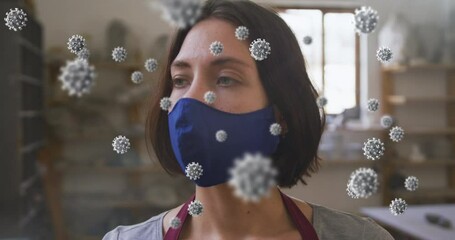 Animation of covid 19 cells over woman in pottery studio wearing face mask - Powered by Adobe