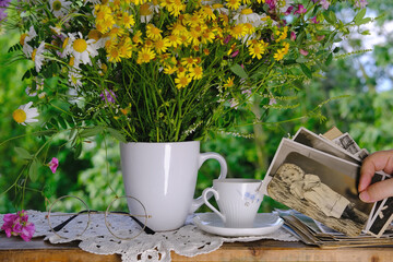 female hands going through old photos of 40s, 50s, bouquet of wildflowers, handmade lacy, cup of tea on table in garden, concept of genealogy, memory of ancestors, family tree, family archive