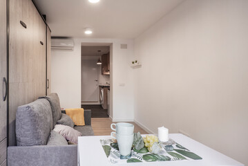 white folding dining table with cups, folding sofa bed with cushions and open kitchen in the background