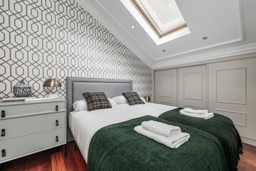 Bedroom with double bed with green cushions and blankets, white towels, built-in wardrobe with sliding doors, pale green chest of drawers in attic room