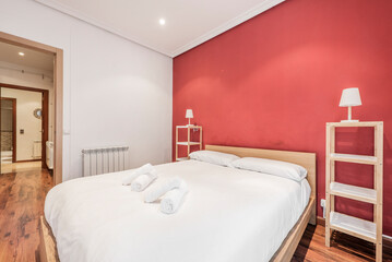 Bedroom with king size bed with wooden headboard, twin wooden shelves as side tables, white aluminum radiator and red painted wall with chestnut wood flooring