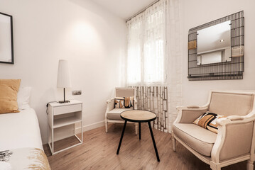 Bedroom with king size bed with white metal and wood nightstands and reading area with two armchairs with wooden armrests and mirror with black wire notes