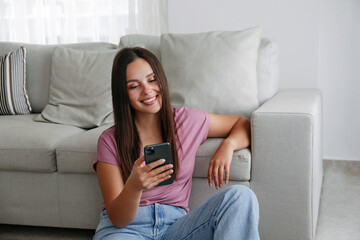 Portrait of young brunette woman wearing mom jeans sitting on the floor leaning on the couch texting and smiling. Joyful female having fun video phone call. Background, copy space, close up.