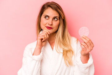 Young caucasian woman holding facial sponge isolated on pink background looking sideways with doubtful and skeptical expression.