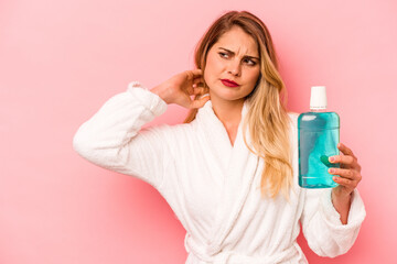 Young caucasian woman holding mouthwash wearing bathrobe isolated on pink background touching back of head, thinking and making a choice.