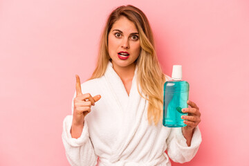 Young caucasian woman holding mouthwash wearing bathrobe isolated on pink background having some great idea, concept of creativity.