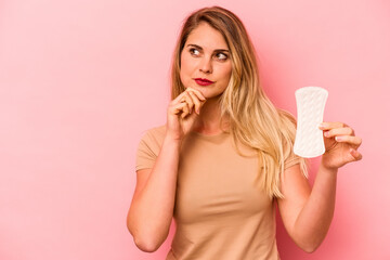 Young caucasian woman holding sanitary napkin isolated on pink background looking sideways with doubtful and skeptical expression.