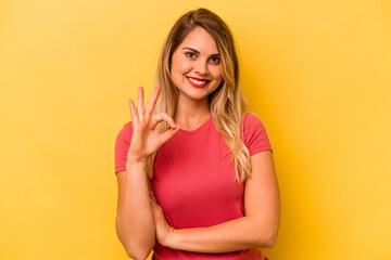 Young caucasian woman isolated on yellow background winks an eye and holds an okay gesture with hand.