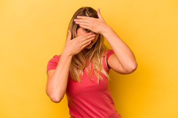 Young caucasian woman isolated on yellow background blink at the camera through fingers, embarrassed covering face.