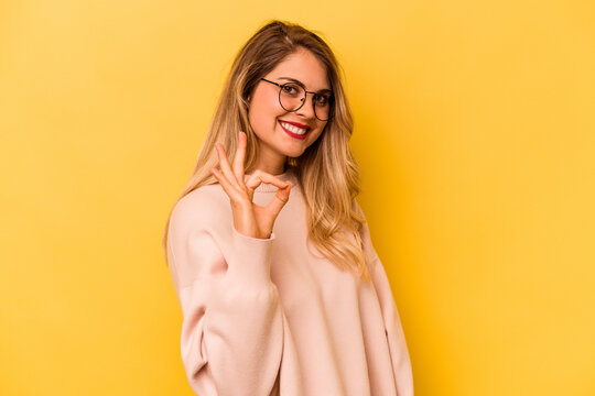 Young caucasian woman isolated on yellow background winks an eye and holds an okay gesture with hand.