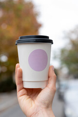 Young female hand holds paper cup of coffee, take away. Tree background