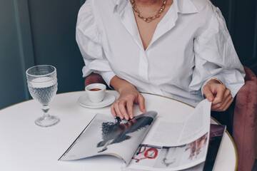 Young stylish woman drinking coffee at cafe, while reading magazine