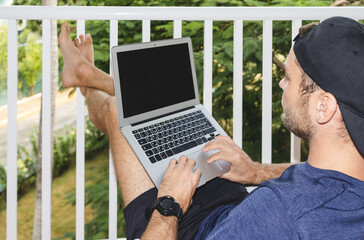 Young man freelancer in cap sitting on balcony with crossed feet and using his laptop, male working on laptop, rear view.