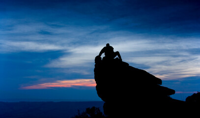 unidentified man sitting on a rock outcrop in silhouette with deep blue and pink evening sky behind