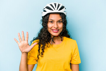 Young hispanic woman wearing a helmet bike isolated on blue background smiling cheerful showing number five with fingers.