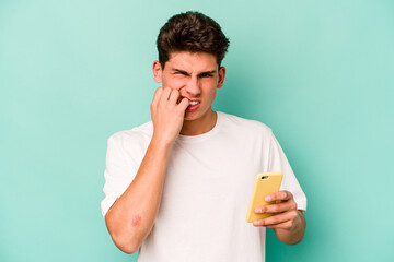 Young caucasian man holding mobile phone isolated on blue background biting fingernails, nervous and very anxious.