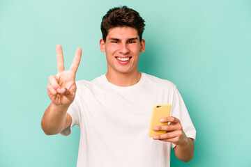 Young caucasian man holding mobile phone isolated on blue background showing number two with fingers.