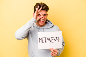 Young caucasian man holding metaverse placard isolated on yellow background excited keeping ok gesture on eye.