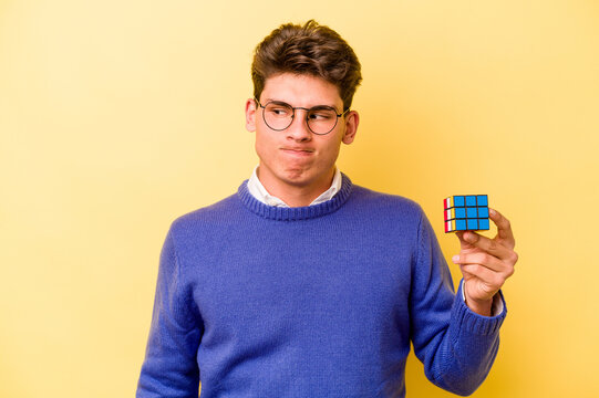 Young Caucasian Man Holding A Rubiks Cube Isolated On Yellow Background Confused, Feels Doubtful And Unsure.