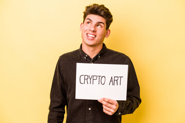 Young caucasian man holding crypto art placard isolated on yellow background dreaming of achieving goals and purposes