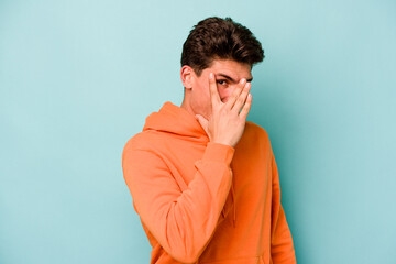 Young caucasian man isolated on blue background blink at the camera through fingers, embarrassed covering face.