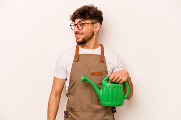 Young gardener hispanic man holding a watering can isolated on white background looks aside smiling, cheerful and pleasant.