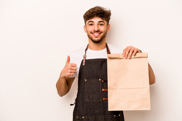 Young hispanic clerk man holding a take away bag isolated on white background smiling and raising thumb up