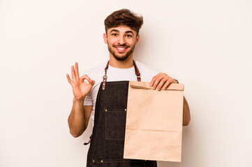 Young hispanic clerk man holding a take away bag isolated on white background cheerful and confident showing ok gesture.