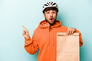 Young hispanic man wearing a helmet bike holding a take away food isolated on blue background pointing to the side