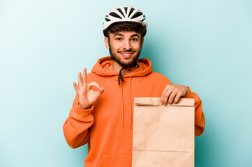 Young hispanic man wearing a helmet bike holding a take away food isolated on blue background cheerful and confident showing ok gesture.