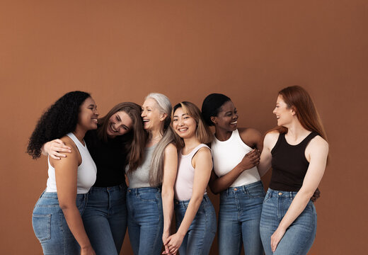 Six laughing women of different body types and ethnicities in studio. Group portrait of smiling females standing together against brown background.