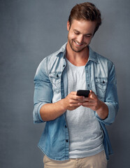 Fototapeta premium Connected to his friends whenever, wherever. Shot of a happy young man using his smartphone while standing against a gray background in the studio.