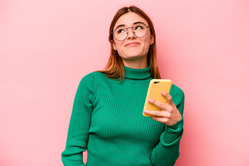 Young caucasian woman holding mobile phone isolated on pink background dreaming of achieving goals...