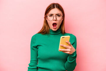 Young caucasian woman holding mobile phone isolated on pink background screaming very angry and...