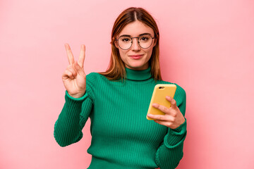 Young caucasian woman holding mobile phone isolated on pink background showing number two with...