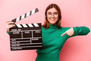 Young caucasian woman holding a clapperboard isolated on pink background person pointing by hand to...