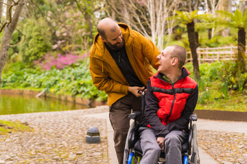 a person with a disability in the wheelchair being pushed by a friend in a public city park, having fun in spring - obrazy, fototapety, plakaty
