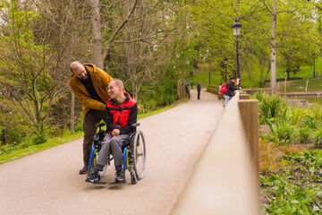 A person with a disability in a wheelchair being pushed by a friend in a public city park, strolling along a path