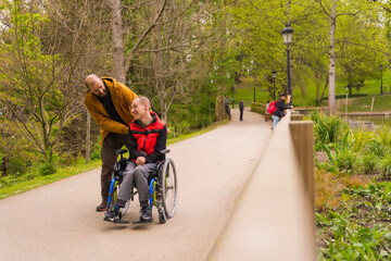 A person with a disability in a wheelchair being pushed by a friend in a public city park,...