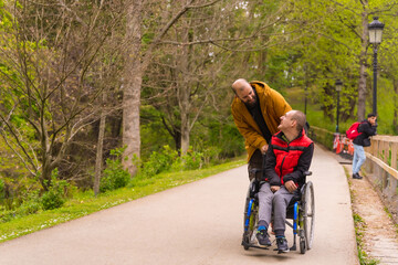 A person with a disability in a wheelchair being pushed by a friend in a public city park, strolling along a path