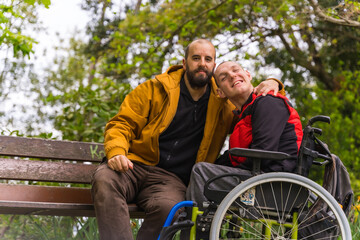 A person with a disability young man in a wheelchair with a friend on a bench in a public park in the city - Powered by Adobe