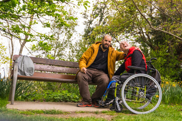 A person with a disability young man in a wheelchair with a friend on a bench in a public park in...