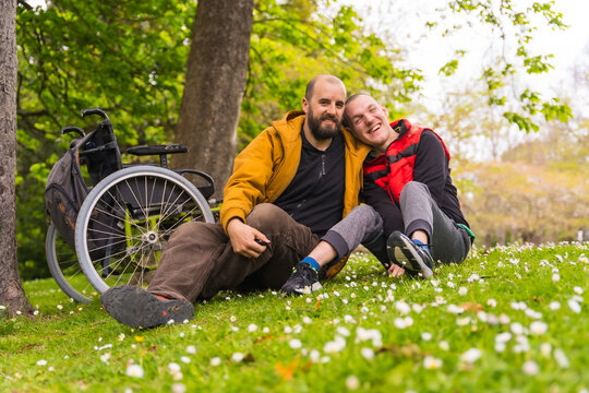 Portrait of a person with a disability with a friend sitting on the grass of a public park in the city, talking and laughing
