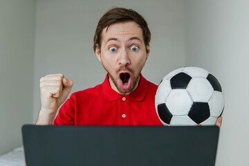Young excited man fan support his football team hold in hand soccer ball watch tv live stream on...