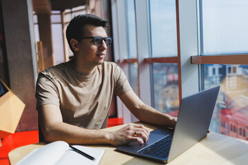 Young handsome smiling smart freelancer in glasses works at a laptop while spending time in a cafe. A positive student sits at a wooden table in a cafe. Happy man sitting while working indoors