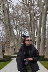 Fototapeta premium Young beautiful woman with camera at park. She has sun glasses. Warm clothes at winter season. Fashion look.