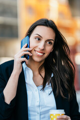 Casually dressed business woman standing on city street and using her smart phone to communicate with someone while holding coffee to go in other hand. Bright sunny day.